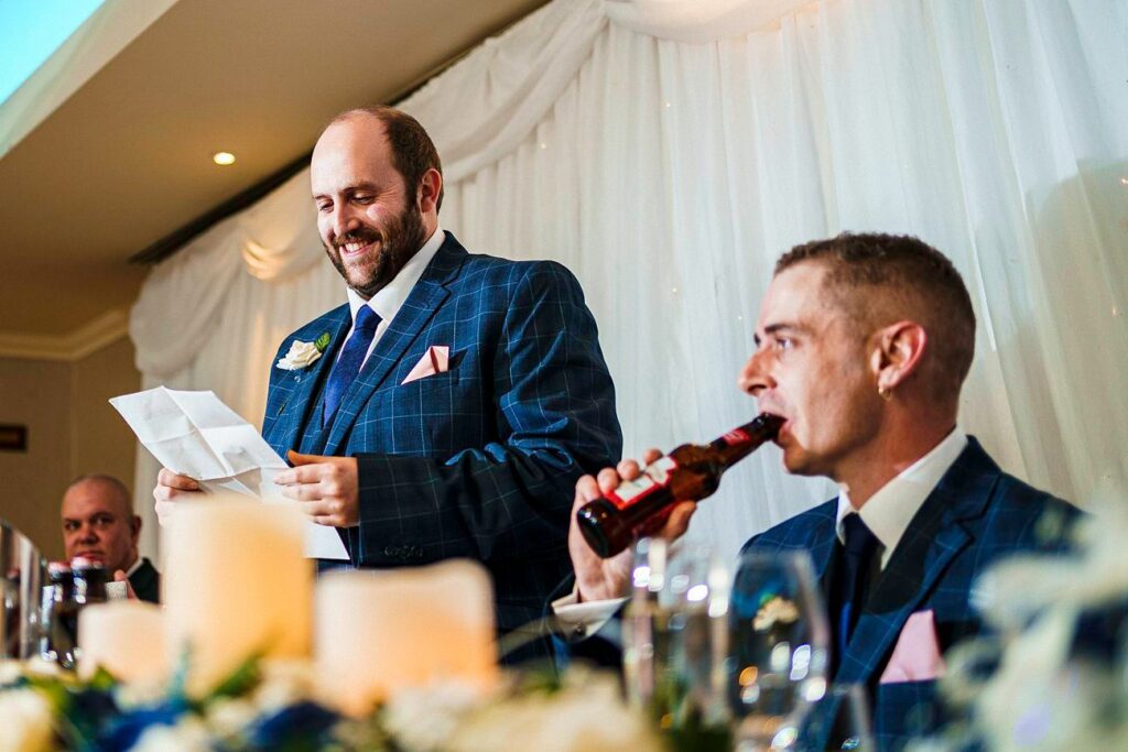 A man in a blue plaid suit stands and smiles while reading from a piece of paper at a wedding reception. Another man seated beside him, also in a blue plaid suit, drinks from a bottle of beer. The table is decorated with candles and flowers. Image by Documentary Wedding Photographer Lewis Bishop Photography.