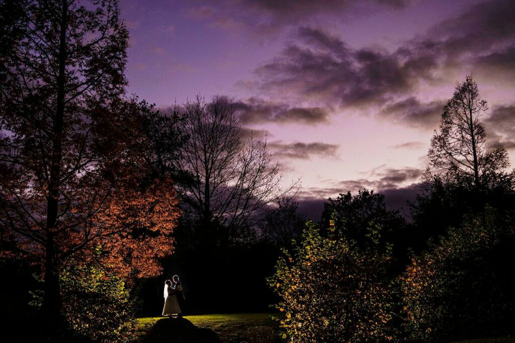 A couple stands in an embrace under a twilight sky with purple and pink hues. Surrounding them are trees with autumn foliage. The couple is centrally illuminated, contrasting with the darker landscape and silhouetted trees, creating a romantic scene. Image by Documentary Wedding Photographer Lewis Bishop Photography.