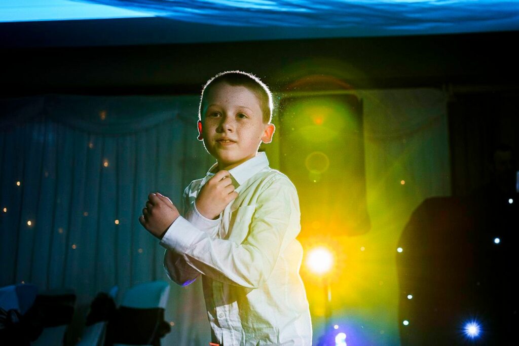 A young boy in a white shirt is standing under colorful stage lighting, with various lights creating a vibrant atmosphere around him. He appears to be looking slightly to his left and is positioned indoors with draped curtains in the background. Image by Documentary Wedding Photographer Lewis Bishop Photography.