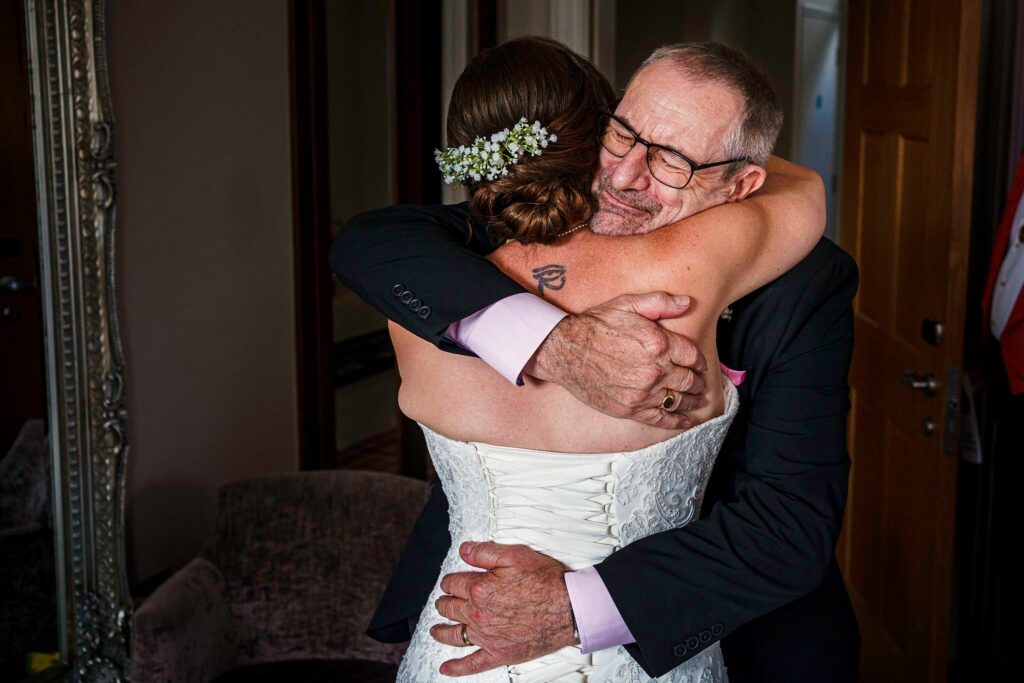 A bride in a white dress and floral headpiece shares an emotional hug with an elderly man wearing glasses and a black suit. They embrace warmly in a room with elegant decor. The bride's back is to the camera, and the man has his eyes closed, showing a tender moment. Image by Documentary Wedding Photographer Lewis Bishop Photography.