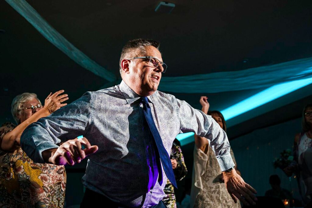 A man wearing a patterned shirt and tie dances energetically at a party. He appears joyful and is surrounded by other people who are also enjoying the festive atmosphere. Blue lighting and draped fabric add to the celebratory mood of the scene. Image by Documentary Wedding Photographer Lewis Bishop Photography.