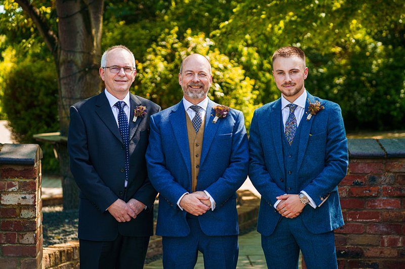 Three men in suits stand together outdoors, posing for a photo. The two older men are on the left and center, and a younger man is on the right. They all wear blue suits with boutonnieres. Image by Documentary Wedding Photographer Lewis Bishop Photography.