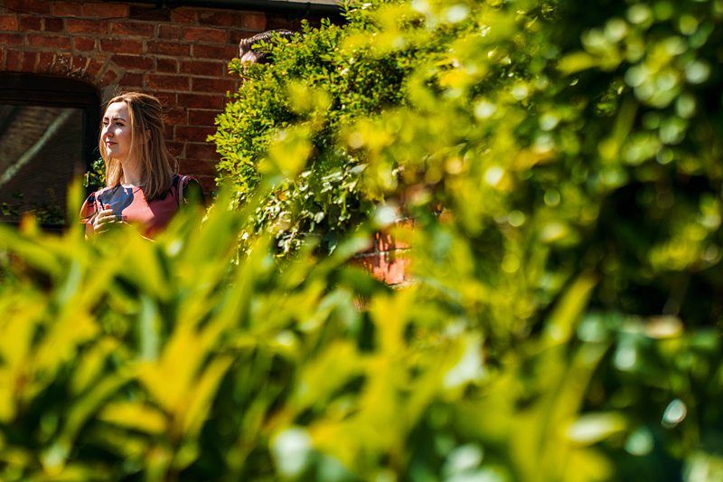 A woman stands outside a brick building, partially obscured by green foliage, holding an object in her right hand. Image by Documentary Wedding Photographer Lewis Bishop Photography.
