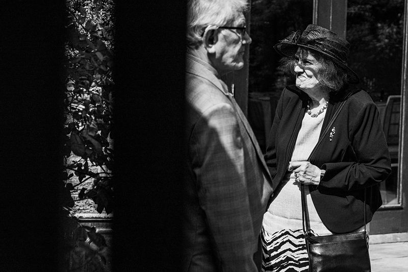 An elderly man and woman are engaged in conversation outside, both dressed in formal attire. The woman is wearing a hat and patterned skirt, holding a handbag. The photo is in black and white. Image by Documentary Wedding Photographer Lewis Bishop Photography.
