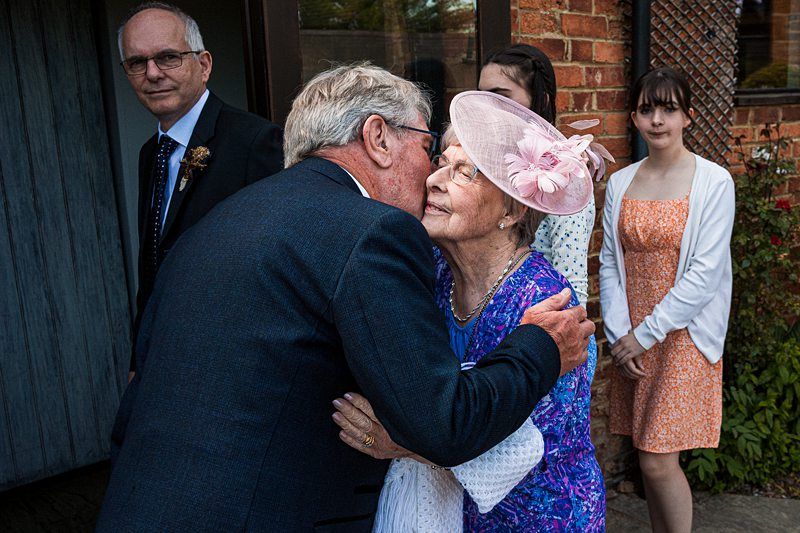 An elderly man hugs an elderly woman dressed in purple with a pink hat. Two younger women and a man stand in the background, observing the scene. The setting appears to be outdoors near a brick building. Image by Documentary Wedding Photographer Lewis Bishop Photography.