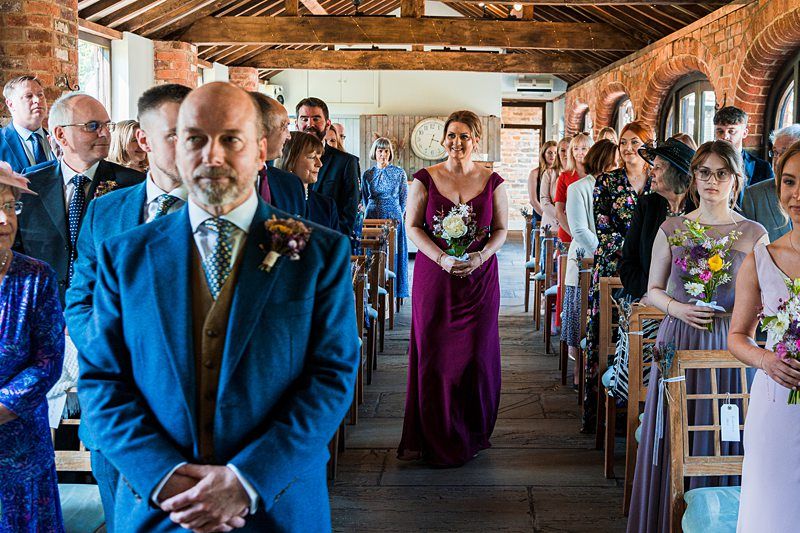 A bride walks down the aisle in a rustic venue, holding a bouquet, while wedding guests stand on both sides. The groom, in a blue suit, stands at the front waiting. Image by Documentary Wedding Photographer Lewis Bishop Photography.