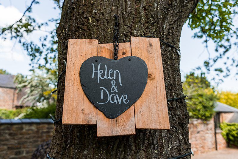 A wooden heart-shaped sign with "Helen & Dave" written on it is attached to a tree trunk outdoors. Image by Documentary Wedding Photographer Lewis Bishop Photography.