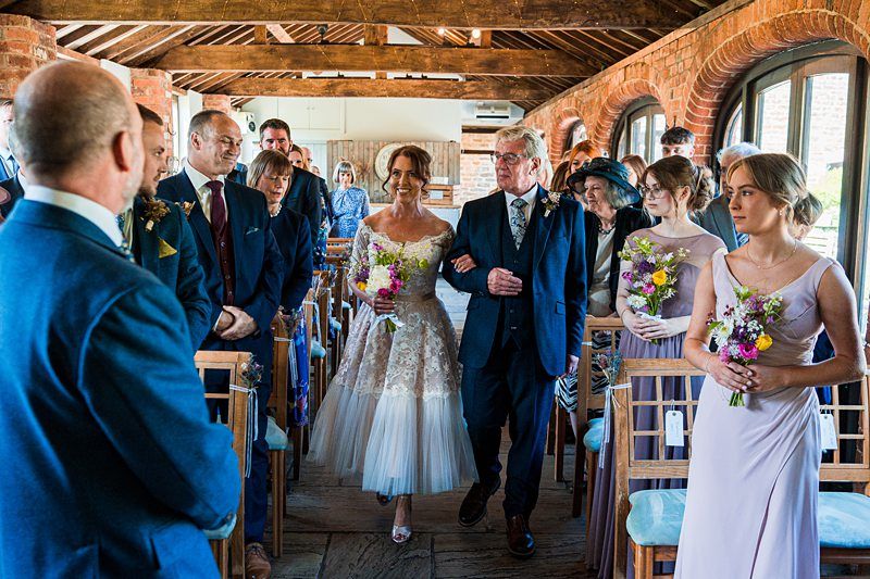 A bride in a white dress walks down the aisle with an older man in a suit, surrounded by guests seated on wooden chairs, inside a rustic venue with exposed beams and brick walls. Image by Documentary Wedding Photographer Lewis Bishop Photography.