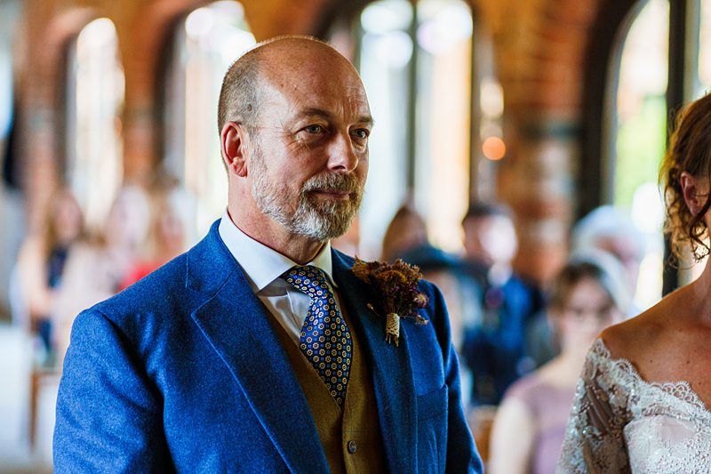 A man in a blue suit with a patterned tie stands indoors, partially facing a woman in a white lace dress. The background is blurred, showing seated guests and arched windows. Image by Documentary Wedding Photographer Lewis Bishop Photography.