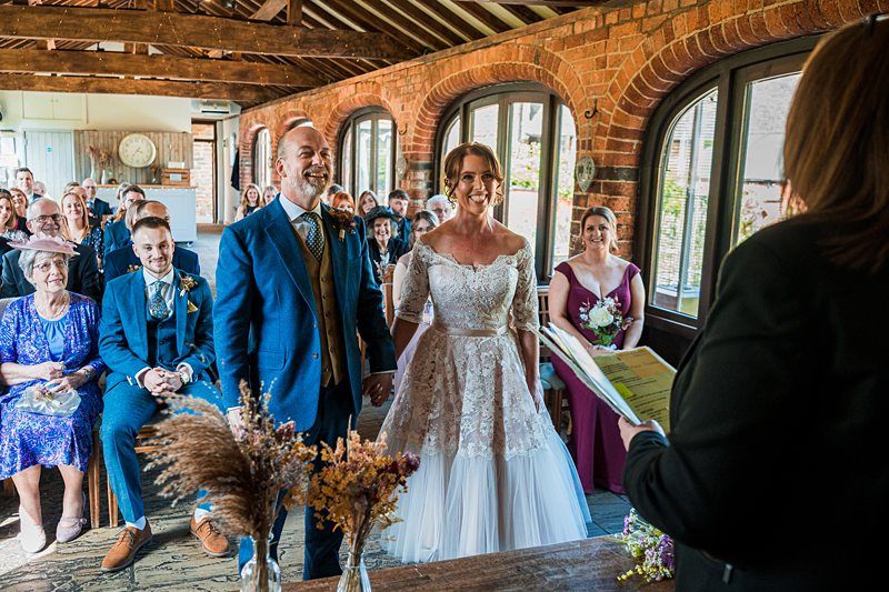 A couple stands before an officiant during a wedding ceremony. Guests are seated in rows behind them in a rustic venue with brick walls and large arched windows. Image by Documentary Wedding Photographer Lewis Bishop Photography.