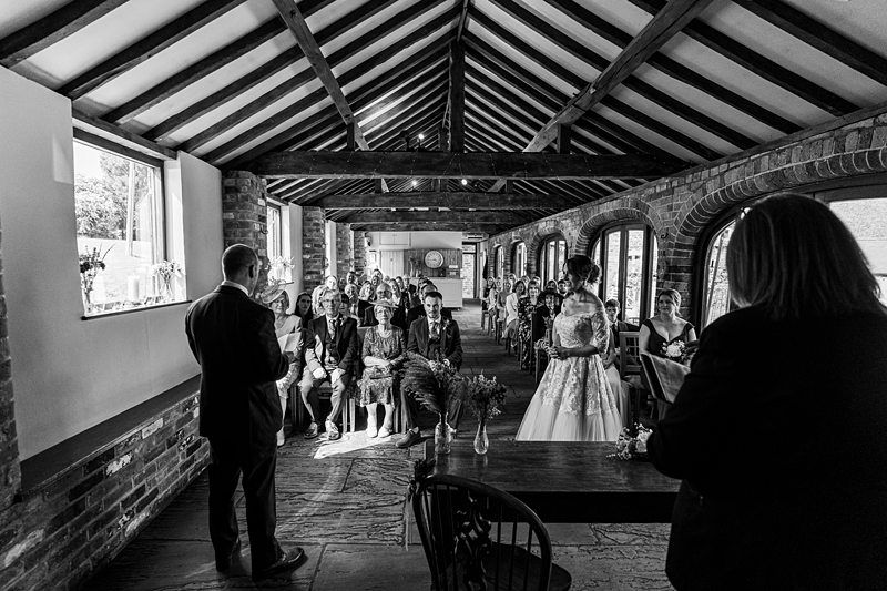 A black and white photo of a wedding ceremony indoors. Guests are seated in rows, and the couple stands at the front with the officiant, under a rustic wooden beamed ceiling. Image by Documentary Wedding Photographer Lewis Bishop Photography.