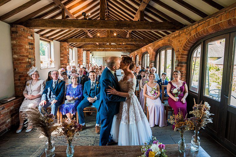 A bride and groom share a kiss at their wedding ceremony in a rustic venue, surrounded by seated family and friends. Image by Documentary Wedding Photographer Lewis Bishop Photography.