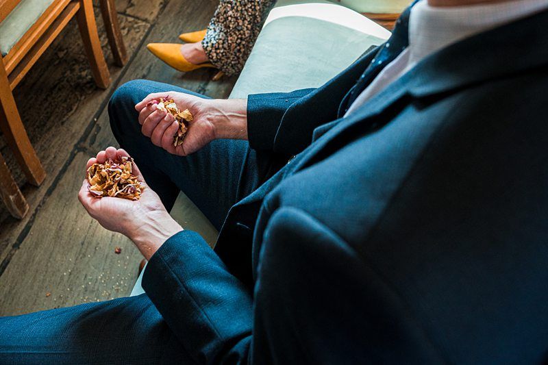A seated individual in formal attire holds dried flower petals in both hands, with a person wearing yellow shoes visible in the background. Image by Documentary Wedding Photographer Lewis Bishop Photography.