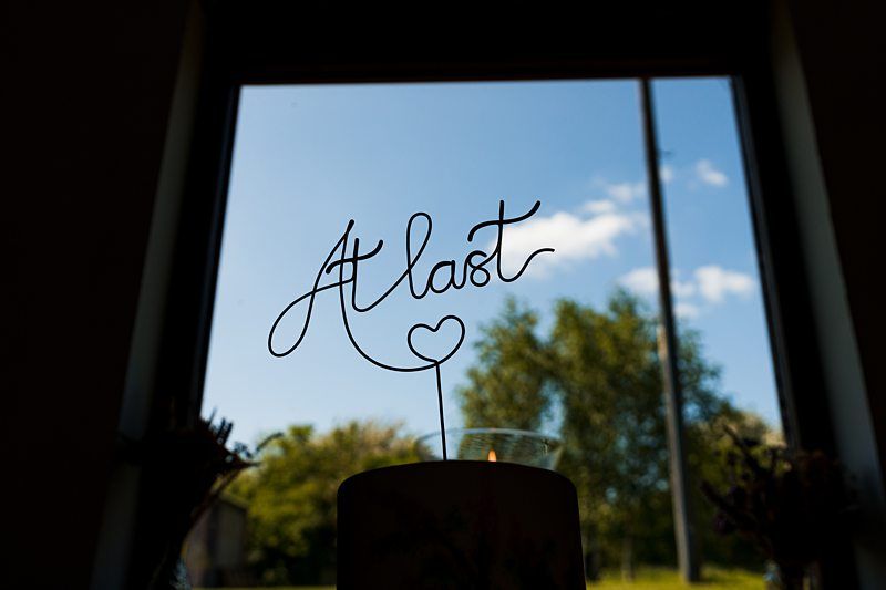 A window provides a backdrop for a decorative wire sign that reads "At last" with a heart shape. The background features a blue sky and green trees. Image by Documentary Wedding Photographer Lewis Bishop Photography.