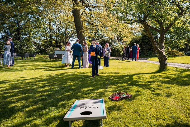 People in formal attire playing lawn games and socializing outdoors on a sunny day, with trees and green grass visible in the background. Image by Documentary Wedding Photographer Lewis Bishop Photography.