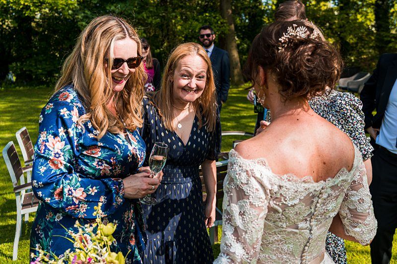 Three women, two holding champagne flutes, converse outdoors in a sunlit garden. One woman wears a white lace dress with an updo hairstyle, while the others wear floral and polka-dot dresses. Image by Documentary Wedding Photographer Lewis Bishop Photography.