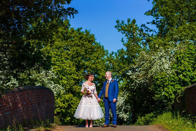 A couple, dressed in wedding attire, holds hands while standing on a brick bridge surrounded by lush greenery under a clear blue sky. The bride wears a white dress, and the groom is in a blue suit. Image by Documentary Wedding Photographer Lewis Bishop Photography.
