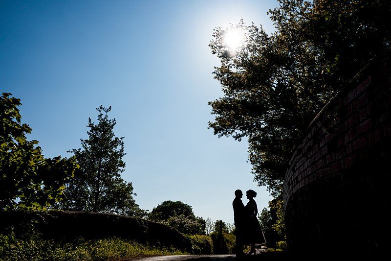 Silhouette of two people standing close together on a path surrounded by trees, with the sun shining brightly in the clear sky above. Image by Documentary Wedding Photographer Lewis Bishop Photography.