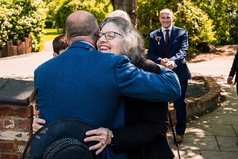 A man in a blue suit hugs a woman with gray hair and glasses outdoors on a sunny day. Another man in a suit smiles in the background. Image by Documentary Wedding Photographer Lewis Bishop Photography.