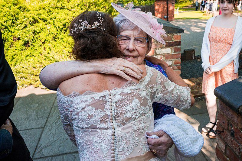 An older woman in a pink hat and glasses warmly embraces a bride in a lace dress outdoors; a few other people are present in the background. Image by Documentary Wedding Photographer Lewis Bishop Photography.