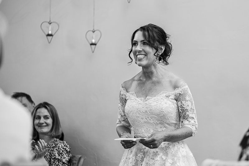 A woman in a lace wedding dress smiles and holds a piece of paper, standing indoors with heart-shaped lanterns in the background and guests applauding. Image by Documentary Wedding Photographer Lewis Bishop Photography.
