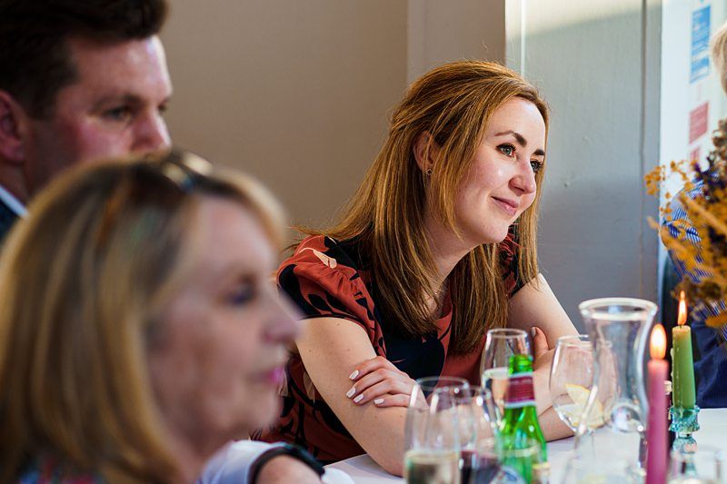 A woman with long brown hair, seated at a table with drinks and flowers, looks attentively to her right. Two other people are partially visible in the foreground and background. Image by Documentary Wedding Photographer Lewis Bishop Photography.