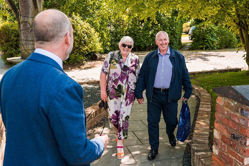 A smiling couple holding hands walk towards a man in a blue jacket. The woman wears a floral dress and sunglasses, while the man wears a striped shirt and jacket. They are outdoors on a sunny day. Image by Documentary Wedding Photographer Lewis Bishop Photography.