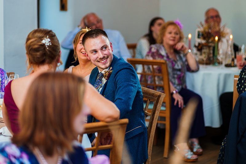 A man in a blue suit is smiling while seated at a table in a room with other people at what appears to be a formal event, such as a wedding or celebration. Image by Documentary Wedding Photographer Lewis Bishop Photography.