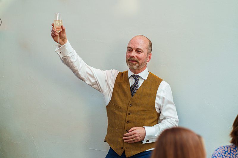 A man in a brown vest and white shirt holding up a glass of champagne while speaking to a partially visible audience. Image by Documentary Wedding Photographer Lewis Bishop Photography.