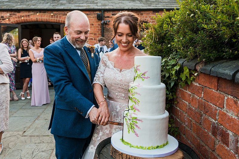 A couple, dressed in formal attire, cuts a three-tiered wedding cake decorated with floral designs outside a brick building, while guests look on in the background. Image by Documentary Wedding Photographer Lewis Bishop Photography.