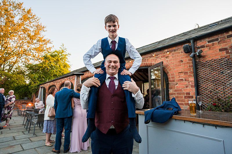 A man wearing a suit holds a boy on his shoulders outside a building with other people in the background. Image by Documentary Wedding Photographer Lewis Bishop Photography.