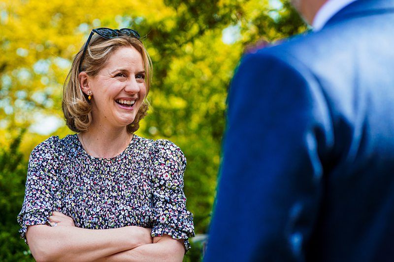 A woman wearing a floral blouse and sunglasses on her head smiles and crosses her arms while conversing with a person in a blue suit. They are outdoors with green foliage in the background. Image by Documentary Wedding Photographer Lewis Bishop Photography.