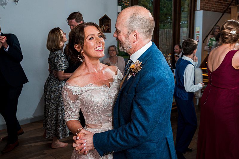 A couple, dressed in formal attire, dance together at an indoor event. The woman wears a white lace dress and the man has a blue suit. Several other attendees are in the background. Image by Documentary Wedding Photographer Lewis Bishop Photography.