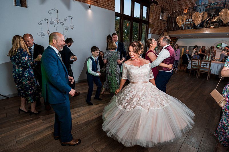 A bride in a white lace gown dances with guests at a wedding reception in a rustic venue with wooden floors and brick walls. Other attendees are seen dancing and socializing in the background. Image by Documentary Wedding Photographer Lewis Bishop Photography.