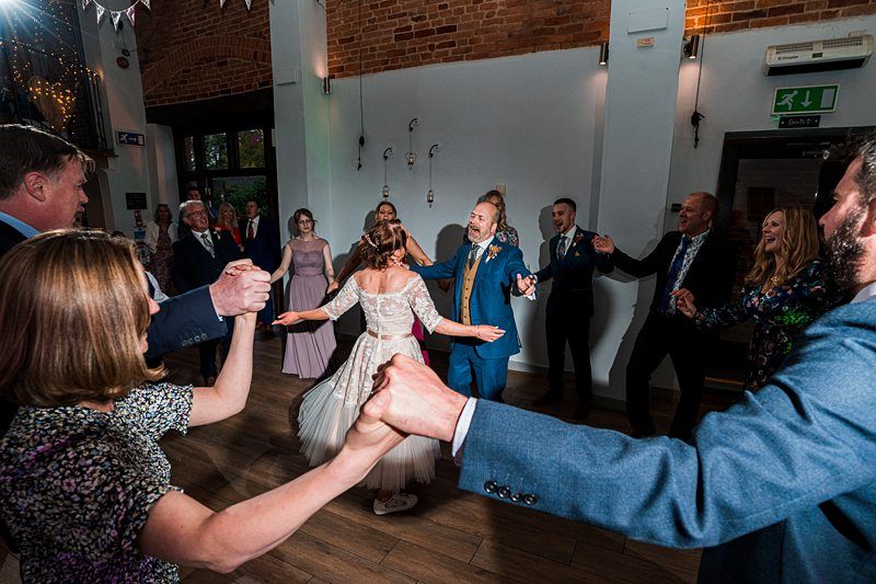 A group of people in formal attire hold hands and dance in a circle at an indoor event with brick walls and wooden flooring. Image by Documentary Wedding Photographer Lewis Bishop Photography.