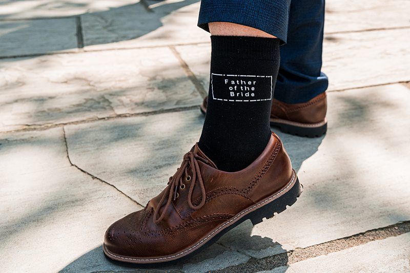 A person wearing brown leather dress shoes and black socks, which have "Father of the Bride" written on them, is standing on a paved surface. Image by Documentary Wedding Photographer Lewis Bishop Photography.