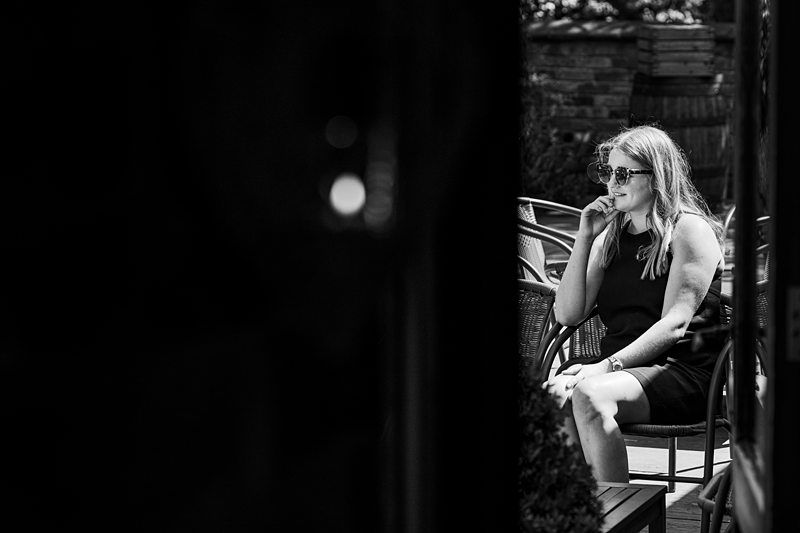 A woman sits alone on an outdoor chair in the shade, wearing sunglasses and a black dress, with a contemplative expression. Image by Documentary Wedding Photographer Lewis Bishop Photography.