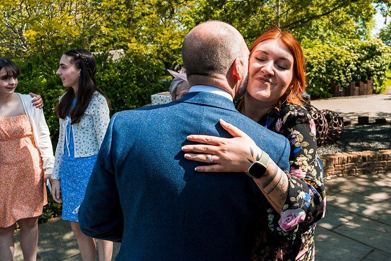 A bald man in a blue suit embraces a woman with brown hair and a floral dress. Three women in summer dresses stand in the background. The setting appears to be an outdoor garden. Image by Documentary Wedding Photographer Lewis Bishop Photography.