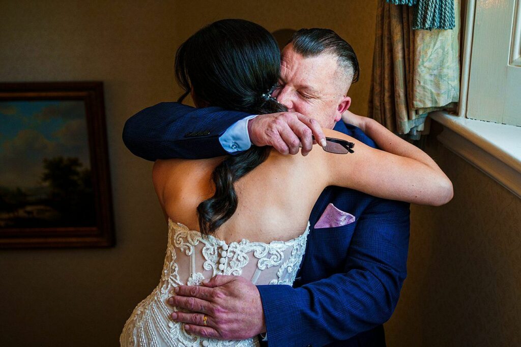 A bride in a white lace dress hugs a man in a navy suit. The man has his eyes closed and appears emotional as he tightly embraces her. They are indoors, near a window with drapes, and a framed painting hangs on the wall in the background. Image by Documentary Wedding Photographer Lewis Bishop Photography.