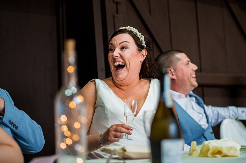 A bride and groom laugh heartily while seated at a table, surrounded by bottles and glassware during a wedding reception. Image by Documentary Wedding Photographer Lewis Bishop Photography.