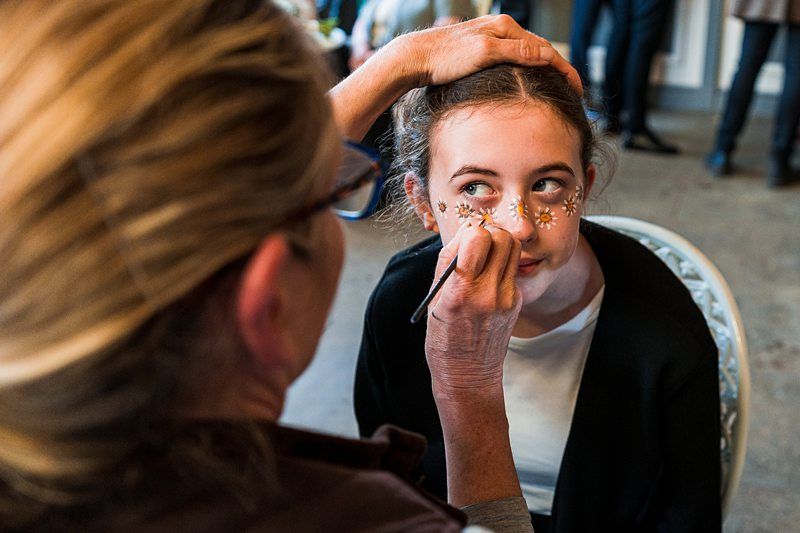 A girl is having her face painted with flower designs by an artist in a casual indoor setting. Image by Documentary Wedding Photographer Lewis Bishop Photography.