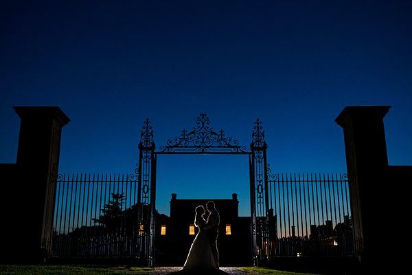 A silhouette of a couple stands closely together under an ornate gate against a deep blue twilight sky. Warm lights from a distant building illuminate behind them, creating a romantic and serene atmosphere. Image by Documentary Wedding Photographer Lewis Bishop Photography.