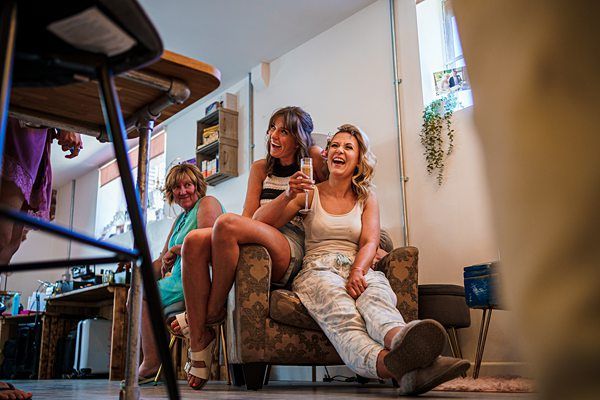 Three women share a laugh in a cozy, well-lit living room. One woman sits on a cushioned chair with a drink in hand, while the others sit close together on the armrest. A wooden chair and a cluttered table are in the foreground, adding a casual vibe to the scene. Image by Documentary Wedding Photographer Lewis Bishop Photography.