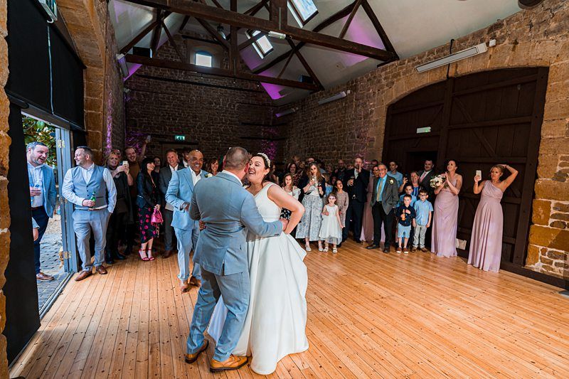A couple shares their first dance in a rustic hall with a stone wall. Guests, including children and adults in formal attire, watch and smile. Image by Documentary Wedding Photographer Lewis Bishop Photography.