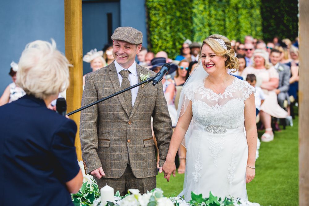 A couple stands holding hands, smiling broadly during an outdoor wedding ceremony. The groom, in a brown plaid suit and flat cap, and the bride, in a white lace dress and veil, face the officiant. Seated guests watch the event, with greenery and a blue wall as the backdrop. Image by Documentary Wedding Photographer Lewis Bishop Photography.