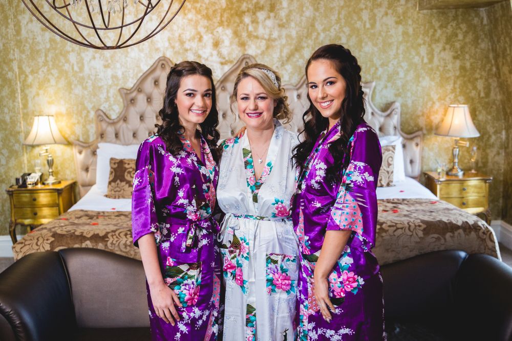Three women are standing and smiling in a bedroom. The woman in the center is wearing a white floral robe, while the women on either side are wearing matching purple floral robes. The room is elegantly decorated with a plush bed, nightstands, and lamps in the background. Image by Documentary Wedding Photographer Lewis Bishop Photography.