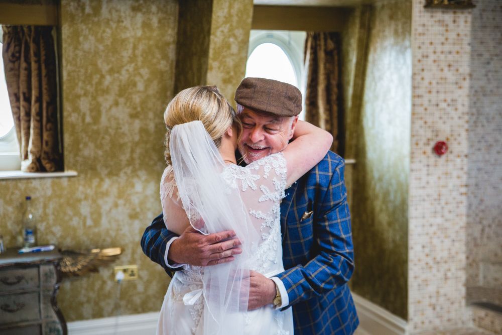 A bride in a white wedding dress and veil hugs an elderly man in a blue plaid suit and flat cap with eyes closed and a joyful expression. The background features golden textured walls and a small window with curtains. Image by Documentary Wedding Photographer Lewis Bishop Photography.