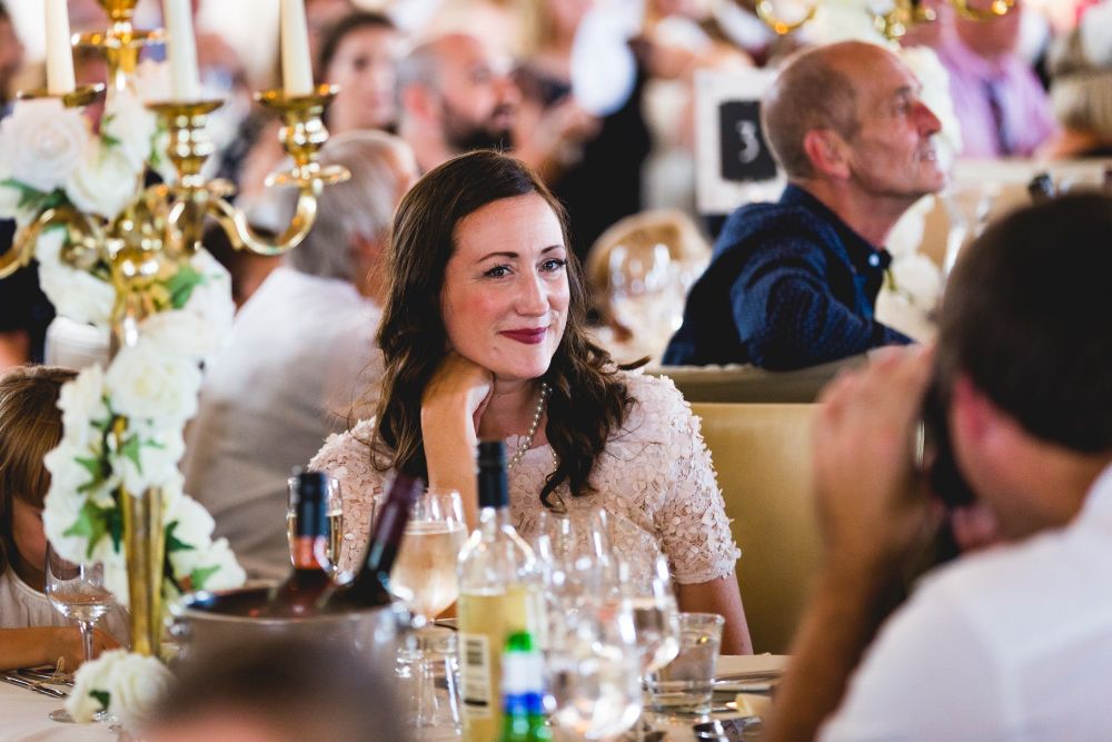 A woman with long, dark hair and wearing a light-colored top is sitting at a table during a formal event. She is smiling and resting her chin on her hand. The table is adorned with wine bottles, glasses, and floral arrangements. Other people are seated in the background. Image by Documentary Wedding Photographer Lewis Bishop Photography.