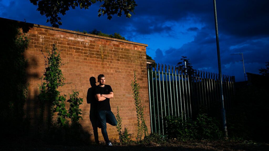 A man in a black shirt and jeans stands against a brick wall with arms crossed. It's dusk with a deep blue sky. The wall has some green plants growing on it, and there's a tall metal fence beside him. The scene is partially shaded by a tree. Image by Documentary Wedding Photographer Lewis Bishop Photography.