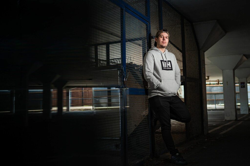 A person in a light gray hoodie and black pants leans against a metal fence in a dimly lit parking garage. The individual looks relaxed with hands in pockets and the background includes structural columns and partial sunlight. Image by Documentary Wedding Photographer Lewis Bishop Photography.